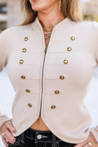 Close-up shot highlighting the zipper closure and double-breasted antique gold buttons on a beige fine-knit jacket. Features textured horizontal ribbing across the front bodice, a raised funnel neckline, and the top waistband of black velvet trousers.