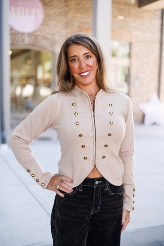 Woman posing outdoors in a beige napolean-style zip-front cardigan jacket featuring double-breasted antique brass button accents and textured horizontal ribbing across the chest. The jacket features a high collar, cuff button details, and a curved tulip hem resting at the waist of high-waisted black pants with white contrast stitching. She stands smiling with hands on her hips, styled with gold jewelry in front of a storefront background.