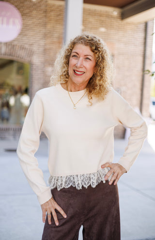 Front-facing mid-shot of a woman modeling a long-sleeve cream fine-knit top with scallop-edged lace layering along the bottom hem. She stands with one hand resting at her hip, highlighting the contrast between the cream lace detail and dark brown suede pants.
