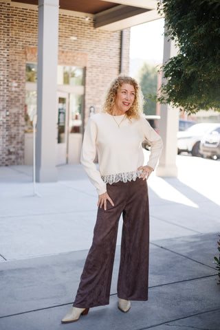 Woman posing outdoors in a long-sleeve cream sweater featured with delicate white eyelash lace hem trim. The top is paired with high-waisted dark brown faux-suede wide-leg pants and beige pointed-toe boots, styled in front of an urban brick building backdrop.