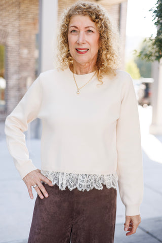 Three-quarter front portrait of a smiling woman wearing a cream crewneck sweater accented with intricate floral lace peplum-style trim at the waist. Paired with chocolate brown wide-leg suede trousers and a dainty gold pendant chain necklace.