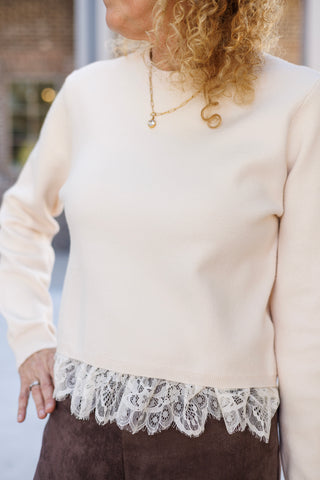 Close-up shot focusing on the hem and texture of a cream crewneck knit top accented with a delicate white floral lace trim. Detailed styling highlights the layered gold link necklace with a round crystal pendant against the soft sweater fabric