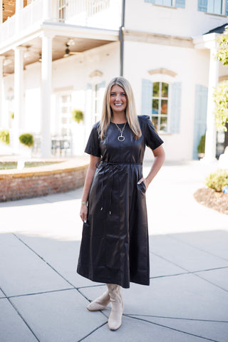 Woman posing outdoors in a dark espresso brown faux-leather short-sleeve midi dress featuring a crew neckline, tailored panel stitching, and a thin tie-belt waist. Styled with tall beige suede boots, a gold chain necklace with an equestrian stirrup pendant, and hands resting near side pockets against a bright architectural background
