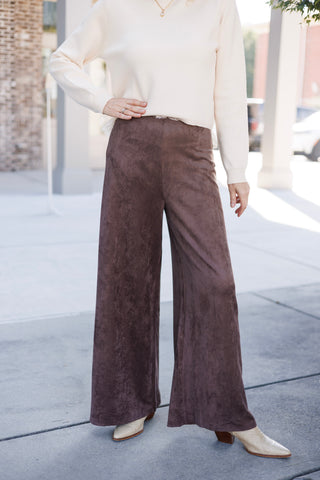 A mid-shot highlighting the soft texture and tailored high-waisted fit of the chocolate brown faux-suede wide-leg pants. Styled with a cream sweater resting right above the smooth waistband and beige textured boots peeking out from beneath the full-length wide leg openings.