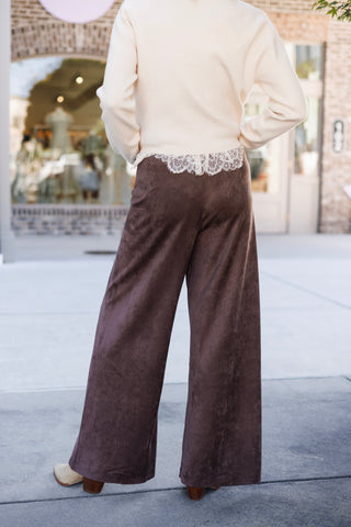 Back view of the chocolate brown faux-suede wide-leg pants showcasing the clean, unadorned back panel design and comfortable stretch drape. Styled under a long-sleeve cream sweater with white lace trim peeking out along the back waist.