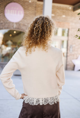 Back view of a woman wearing a cream long-sleeve knit sweater with a white lace layer peeking below the hem, paired with dark brown suede pants. She stands with her back turned to reveal the smooth back panel construction and fine ribbed cuffs.