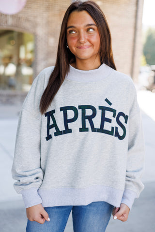 Upper body shot showcasing the relaxed silhouette of the light grey mock-neck pullover sweatshirt. Highlights the collegiate-style navy and hunter green plaid block lettering spelling "APRÈS" stitched across the chest, styled with long dark hair worn down.