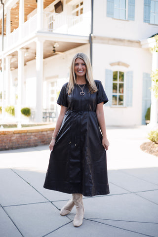 Full-length view of a smiling woman holding out the sides of an A-line dark brown faux-leather midi dress. The short-sleeve dress features structured vertical seams across the bodice and skirt, cinched at the waist with a thin self-tie drawstring belt with metal aglets.