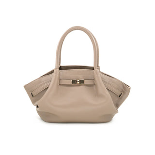 Front view of a chic taupe-beige textured leather tote bag set against a crisp white background. The handbag features a wide, uniquely flared trapezoid silhouette with softly gathered side gussets and two structured rolled top handles. The front is accented with a horizontal leather belt strap secured by a rectangular, polished gold-tone turn-lock closure detail in the center.