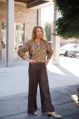 Woman standing outdoors on a sidewalk posing with hands on hips in a retro-inspired floral print blouse featuring warm earth tones of orange, teal, cream, and brown. The blouse features elasticated three-quarter blouson sleeves, light blue braided trim along the raglan seams and front placket, and a split Henley button neck. Tucked into high-waisted dark chocolate brown suede wide-leg trousers, styled with pointed cream western boots and a gold link chain necklace.