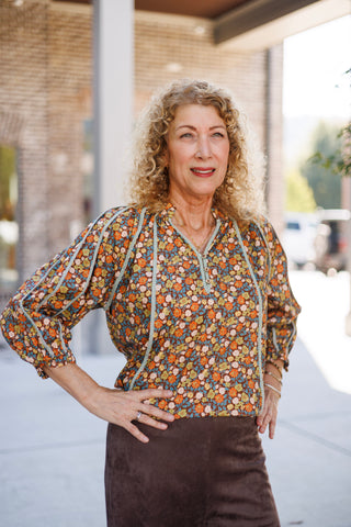 Close-up portrait view of the vintage floral peasant blouse highlighting the intricate micro-floral pattern in shades of marigold, rust, teal, and brown. Features a split V-neckline with a henley button placket, braided trim framing the neckline and raglan shoulder seams, styled with a delicate gold chain pendant necklace.