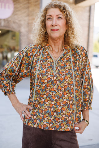 Mid-length front shot showcasing the bohemian floral blouse with light blue contrast braided trim defining the vertical bodice panels and raglan sleeves. Features a partial button front henley placket, blouson three-quarter sleeves with gathered cuffs, and a relaxed silhouette worn tucked into dark brown suede pants.