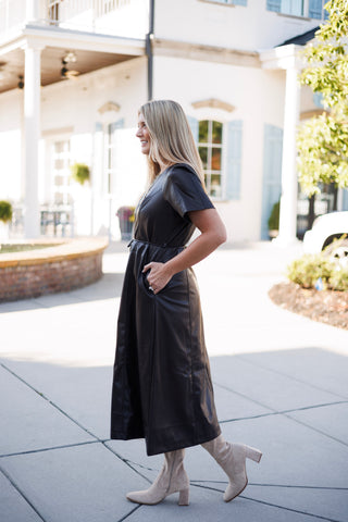 Side-profile view of a woman modeling a dark brown faux-leather midi dress with her hand tucked into a functional side pocket. Highlights the short sleeve design, A-line skirt drape, and knee-high beige suede block-heel boots.