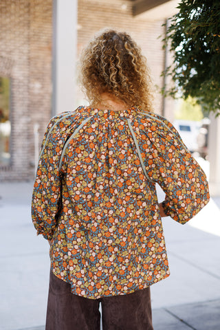 Back view from the waist up showing the continuous small-scale floral print across the upper back, gathered neck seam line, and contrast braided trim framing the raglan sleeve shoulders. Styled with curly blonde hair worn down loose.