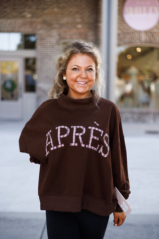 Close-up shot focusing on the upper body and texture of the brown mock-neck pullover, showing the detailed pink plaid fabric infill and dark brown embroidered outline of the "APRÈS" chest lettering. Complemented by stud earrings and wavy blonde hair