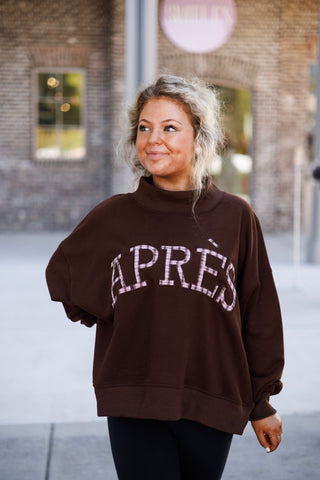 Mid-length portrait view showcasing the relaxed slouched drape of the dark brown fleece pullover top. Highlights the pink plaid block lettering spelling "APRÈS" across the chest, elevated mock neckline, and cozy drop-shoulder sleeve silhouette.