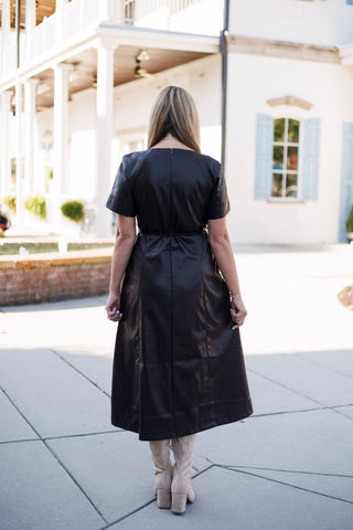 Back view of the dark espresso brown faux-leather midi dress showcasing the center back zipper closure, horizontal waist seam, and tailored back paneling. The model stands with her back turned, showing the modest hem length above knee-high beige boots.