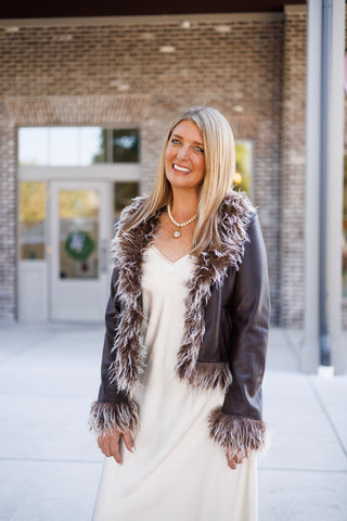 Close-up angled portrait of a smiling woman showing the plush texture of the frosted brown faux-fur collar and wrist cuffs on a fitted dark brown leatherette jacket. Styled over an ivory slip dress with a hand resting near the side pocket.