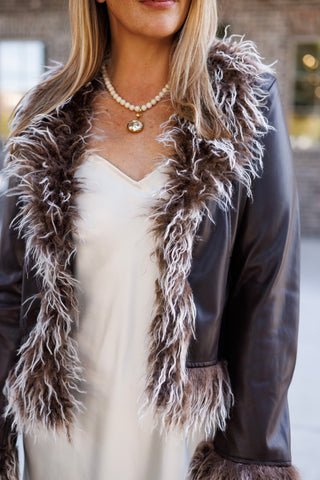 Detail shot focusing on the neckline and lapel construction of an open brown faux-leather outerwear jacket. The jacket features prominent shaggy fur trimming along the lapel edges and sleeve cuffs, layered over a cream satin camisole bodice and beaded necklace.
