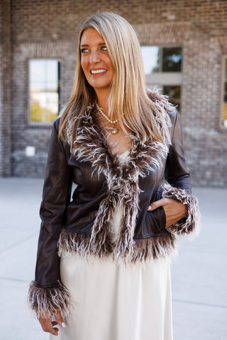 Woman posing outdoors in a dark brown faux-leather jacket featuring fluffy, frosted brown-and-white faux-fur trim along the lapels, cuffs, and hem. Styled open over a flowing cream satin slip dress paired with metallic rose gold pointed-toe pumps and a beaded pendant necklace in front of a brick storefront setting.