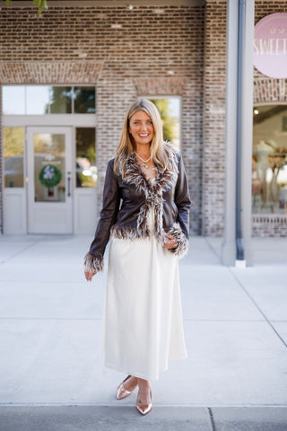 Woman posing outdoors in a dark brown faux-leather jacket featuring fluffy, frosted brown-and-white faux-fur trim along the lapels, cuffs, and hem. Styled open over a flowing cream satin slip dress paired with metallic rose gold pointed-toe pumps and a beaded pendant necklace in front of a brick storefront setting.