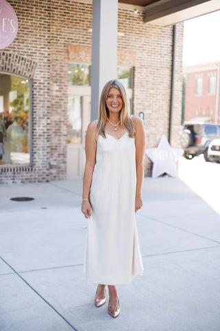 Woman smiling outdoors standing on a sidewalk wearing a fluid champagne-ivory satin slip midi dress featuring a soft V-neckline, adjustable camisole shoulder straps, and a bias-cut skirt drape. Styled elegantly with rose gold metallic pointed-toe pumps, a short pearl strand necklace with a round gold pendant, and gold bracelets in front of a brick storefront.
