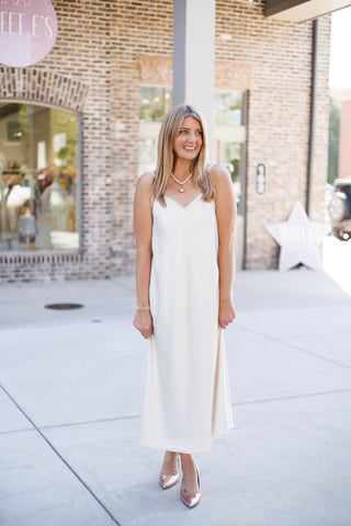 Full-length front view of a woman posing in a sleeveless off-white satin midi slip dress highlighting the relaxed A-line silhouette and subtle shine of the fabric. Styled with metallic pumps and pearl jewelry set against a modern boutique backdrop.