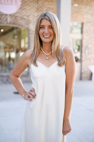 Portrait view focusing on the upper body of the cream satin slip dress, showcasing the V-neck cut, delicate spaghetti straps, and smooth lustrous finish. Accessorized with a classic pearl necklace with a bezel-set pendant and minimal wrist jewelry.