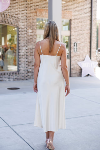 Full-length back view of the champagne satin slip dress showing the straight horizontal back neckline, thin adjustable shoulder straps, and effortless drape down to the midi length hem. Styled with blonde hair worn down and rose gold pointed heels.