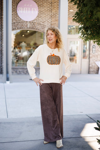 Woman standing outdoors in front of a brick storefront, posing with hands on hips wearing a relaxed cream long-sleeve crewneck sweatshirt. The center chest features a large embroidered leopard print pumpkin graphic outlined with vibrant orange stitching, paired with high-waisted dark chocolate brown suede wide-leg trousers and cream pointed western boots.