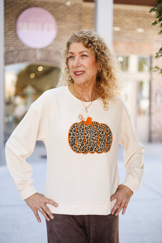 Upper body portrait of a woman wearing the cream long-sleeve graphic pullover sweater with dropped shoulders and ribbed cuffs. Highlights the detailed leopard-print pumpkin appliqué with bright orange embroidery and a curly stem accent, accessorized with a gold chain pendant necklace.