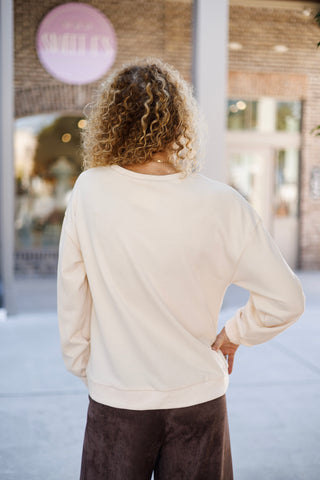 Back view from the waist up showcasing the clean, unprinted design of the cream crewneck pullover, detailing the dropped shoulder seams, soft fleece drape, and ribbed hem band paired with dark brown trousers.