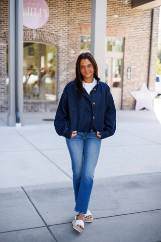 Woman smiling on a sidewalk wearing a dark indigo denim bomber jacket featuring drop shoulders, a high funnel neck collar with button tabs, tortoiseshell buttons over a front zipper, welt hand pockets, and an elastic waist band. Styled casually over a plain white crewneck t-shirt with medium-wash straight-leg jeans and white platform slide sandals.