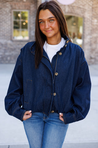 Three-quarter view of the dark wash denim jacket highlighting the oversized utility silhouette, voluminous long sleeves with button cuffs, front button placket with exposed zipper, and contrast amber stitching
