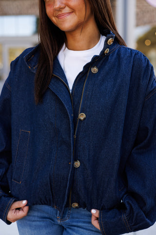 Close-up shot of the dark blue denim bomber jacket displaying the textured denim weave, tortoiseshell button front detail, gold contrast seam stitching, and structured stand-up collar layered over a crisp white tee.