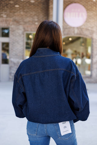 Back view of the dark indigo denim bomber jacket showcasing the stitched back yoke detail, relaxed balloon-style long sleeves, and gathered elastic bottom hem. Styled with slim straight-leg blue denim jeans featuring a brand pocket tag.