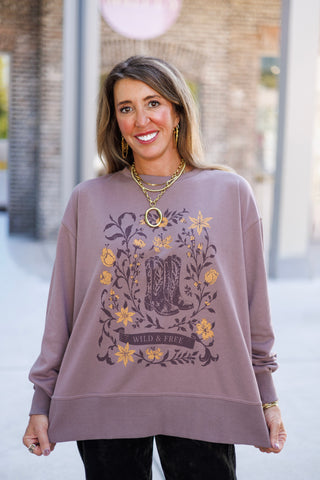 Woman smiling outdoors wearing an oversized mauve-taupe crewneck sweatshirt featuring a dark brown and muted mustard yellow western graphic print. The graphic displays a pair of detailed cowboy boots surrounded by a vintage floral frame and a banner reading "WILD & FREE". Styled casually with dark pants and layered gold chain jewelry.