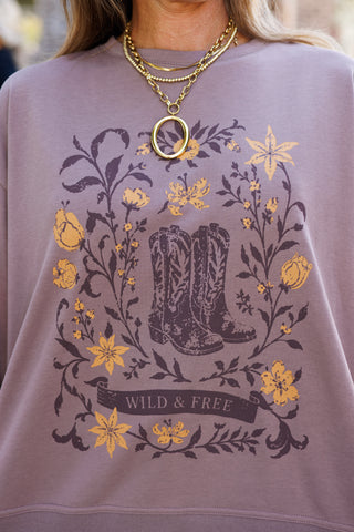 Macro close-up shot focusing on the chest graphic of a dusty mauve crewneck sweatshirt. Highlights the detailed screen print of vintage cowboy boots encircled by wild floral vines, mustard-yellow blossoms, and a bottom banner inscribed with "WILD & FREE". Accented by a gold link chain necklace with a circular pendant.