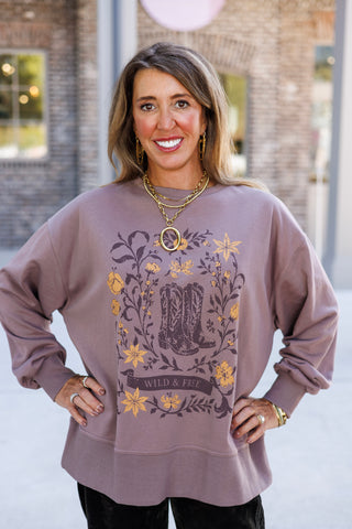 Portrait of a woman posing with hands on hips wearing a soft mauve-brown long-sleeve crewneck sweatshirt with dropped shoulders and a ribbed hem. Center graphic showcases distressed western cowboy boots framed by botanical illustrations and a "WILD & FREE" scroll emblem, paired with a chunky gold ring pendant necklace and gold drop earrings.