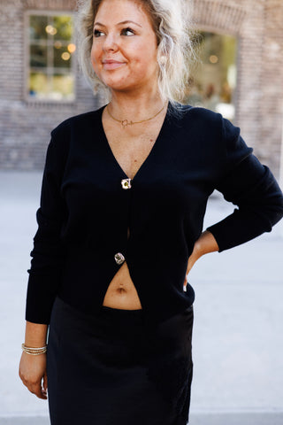 Portrait view highlighting the soft knit texture of the black long-sleeve cardigan top, featuring a deep V-neckline and two textured gold buttons. The top is styled slightly unbuttoned at the hem over a black satin lace-trim skirt, complemented by a delicate gold necklace and beaded bracelets.