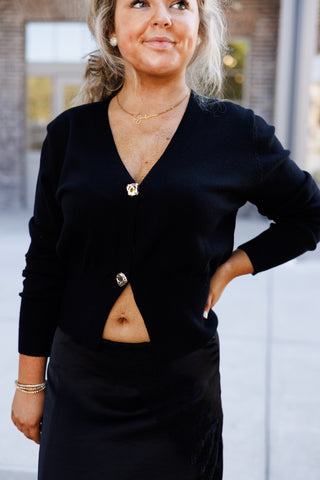 Mid-length front view showcasing the tailored fit of the black knit cardigan sweater with long sleeves, ribbed cuffs, and gold hardware buttons. Tucked subtly over a sleek black satin midi skirt with lace embroidery detailing.
