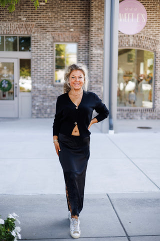 Woman smiling on a sidewalk wearing a chic black long-sleeve V-neck cardigan sweater fastened by two oversized gold statement buttons with an open flared bottom hem. Paired with a high-waisted black satin midi skirt featuring delicate black lace side paneling, styled with casual distress-patterned white sneakers in front of a brick building.