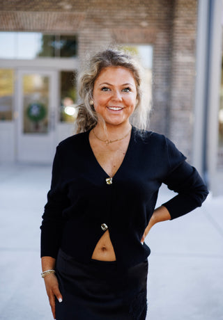Portrait view highlighting the soft knit texture of the black long-sleeve cardigan top, featuring a deep V-neckline and two textured gold buttons. The top is styled slightly unbuttoned at the hem over a black satin lace-trim skirt, complemented by a delicate gold necklace and beaded bracelets