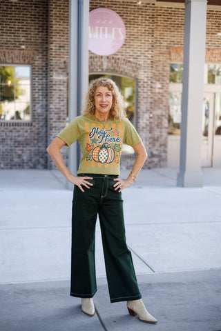 Woman posing outdoors in dark forest green high-waisted wide-leg pants featuring distinct white contrast topstitching along the seams, pockets, and hem. She styles the trousers with an olive green graphic tee reading "Hey There" with pumpkin graphics tucked into the waistband, paired with off-white snakeskin-textured cowboy boots in front of a brick storefront.