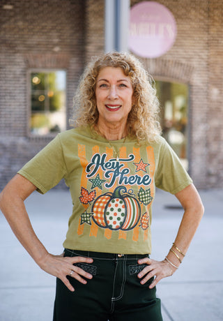 Woman smiling with hands on hips wearing an olive green short-sleeve graphic t-shirt featuring distressed orange vertical stripes, a retro-style pattern-blocked pumpkin, star accents, autumn leaves, and dark green cursive text reading "Hey There". Styled with dark green trousers with contrast stitching and gold jewelry in front of a brick storefront.