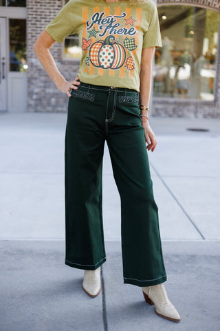 Front view focusing on high-waisted dark green wide-leg trousers detailing the white contrast topstitching, front fly seam, and woven patterned pocket accent trim. Styled with a tucked-in olive fall graphic t-shirt and light beige pointed-toe boots.