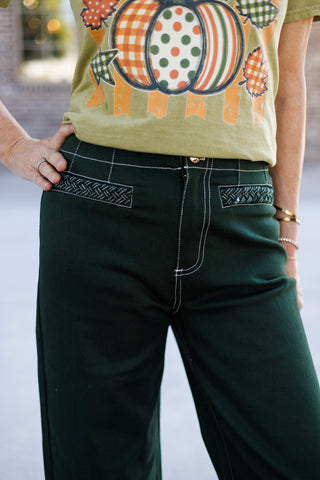 Detailed close-up shot highlighting the structured waistband, copper top button, white contrast stitching, and black-and-white braided trim across the front slash pockets of dark forest green utility-style wide-leg pants.