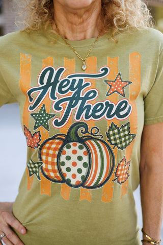 Close-up shot focusing on the front print of the olive green cotton t-shirt. Highlights the detailed screen print featuring dark green "Hey There" script lettering set over weathered orange stripes, a multi-patterned pumpkin in orange, green, and cream tones, gingham stars, and falling leaves.