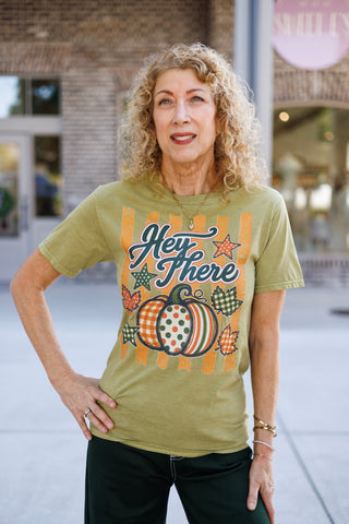 Upper body shot of a woman with curly blonde hair posing in an olive green crewneck graphic tee. The center graphic showcases a colorful pumpkin with polka dot, gingham, and stripe patterns, framed by "Hey There" script typography, autumn leaves, and star illustrations.