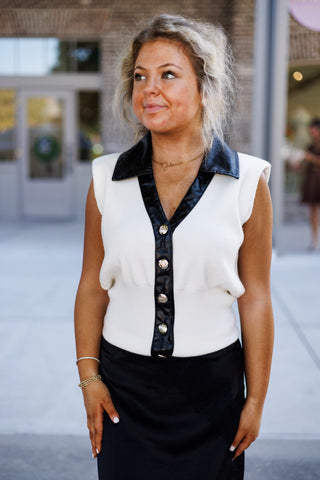 Mid-length front view of a woman posing in a white sleeveless knit top with contrasting black leather-look trim on the collar and button placket. Highlights the metallic gold crest button closures and subtle structured shoulder cap silhouette, styled over a sleek black satin midi skirt.